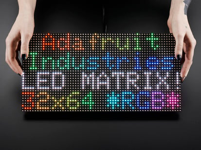64x32 RGB LED Matrix - 6mm pitch - Two white hands hold out an assembled and powered on 64x32 RGB LED Matrix Panel - 6mm pitch. The matrix displays "Adafruit Industries LED MATRIX! 32x64 *RGB*"